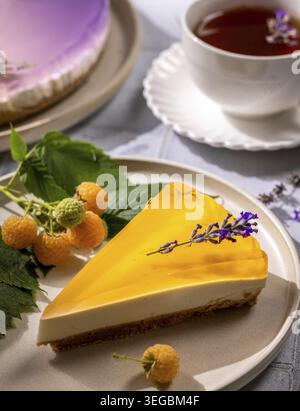 Tranche de cheesecake crémeuse avec garniture de gelée vive, garnie d'une branche de lavande, servie avec des framboises jaunes fraîches et du thé Banque D'Images