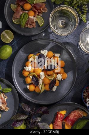 Assiette de départ avec salade de boule de melon avec feta, basilic violet et melon enveloppé de citron vert et jambon servi avec deux verres de vin blanc sur une table sombre Banque D'Images
