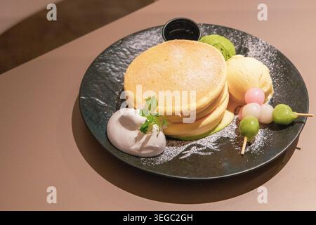 Vue de haut en bas d'une assiette de crêpes de soufflé japonaises avec de la crème glacée matcha, des boules de mochi tricolores et de la crème fouettée sur une assiette sombre, photographiée directement au-dessus dans un café de Kyoto dans une lumière naturelle chaude sur une surface beige neutre. Banque D'Images