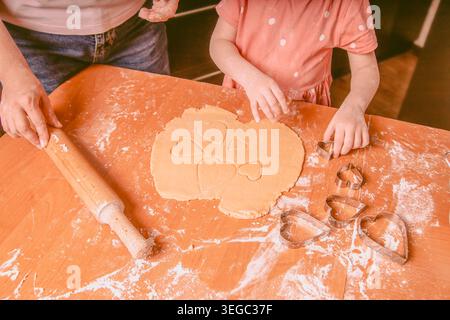 Un adulte et un enfant déroulent la pâte à biscuits sur une table en bois, en utilisant des couteaux en forme de cœur et de la farine dispersée sur toute la surface Banque D'Images