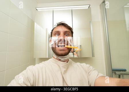 Jeune homme s'amusant avec de la crème à raser sur son visage, tenant un rasoir dans sa bouche Banque D'Images