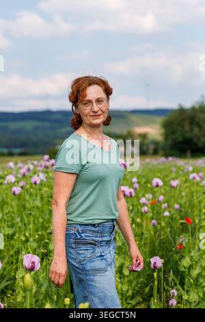 Femme mature avec des lunettes et des cheveux roux bouclés marchant dans un champ de coquelicots rose fleuri et regardant la caméra. Banque D'Images