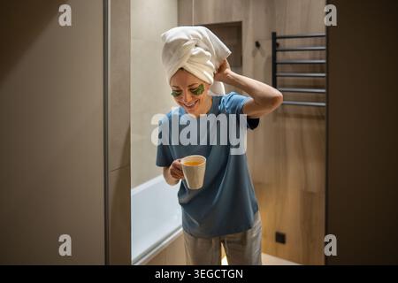 Femme d'âge moyen souriante avec serviette sur la tête tenant une tasse de thé dans l'intérieur moderne de la salle de bains. Rituel de soins personnels le matin ou le soir, style de vie bien-être. Banque D'Images