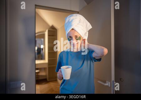 Sereine femme d'âge moyen avec serviette sur la tête et patchs sous les yeux tenant une tasse de tisane regardant la caméra à la maison. Soins personnels le matin ou le soir R Banque D'Images