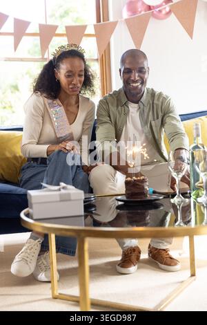 Couple assis sur le canapé du salon, femme portant ceinture et tiare, homme atteignant vers gâteau étincelant Banque D'Images