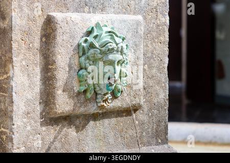 Gros plan d'une fontaine murale en pierre altérée avec un lion patine vert ou un bec verseur mythique, l'eau ruisselant de la bouche à la lumière du jour. Banque D'Images