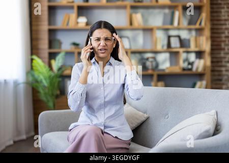 Jeune femme se sentant contrariée et ayant mal à la tête, touchant sa tempe tout en parlant sur un téléphone portable, subissant une conversation stressante ou des nouvelles difficiles dans un cadre de bureau Banque D'Images