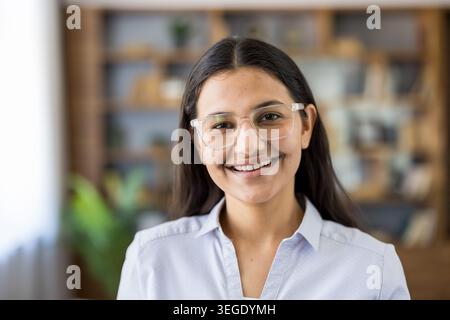 Jeune femme indienne souriante et regardant la caméra, portant des lunettes transparentes et une chemise boutonnée, debout dans un bureau moderne ou un bureau à domicile Banque D'Images