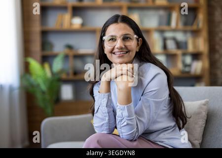 Jeune femme indienne dans des lunettes et chemise bleu clair se détend sur un canapé gris dans un salon moderne lumineux, souriant et regardant loin réfléchi, content et confiant Banque D'Images