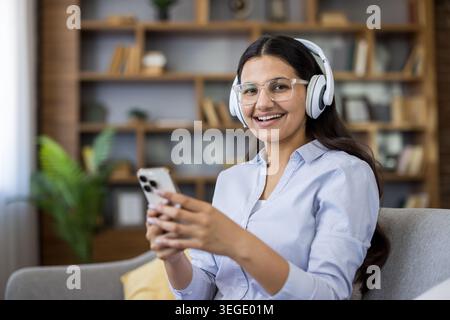 Jeune femme heureuse portant des écouteurs et des lunettes, souriant à la caméra tout en écoutant de la musique ou une leçon en ligne à l'aide d'un smartphone dans un bureau à domicile Banque D'Images