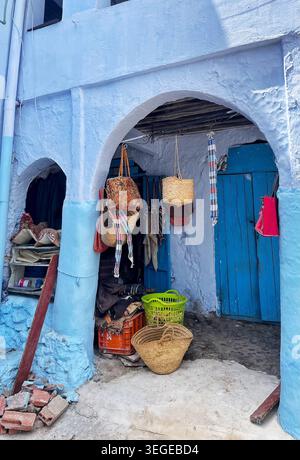 Cour traditionnelle peinte en bleu avec paniers tissés suspendus et sacs colorés faits à la main, 3 août 2025, Chefchaouen, Maroc. Banque D'Images