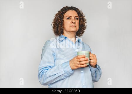 Femme d'âge mûr réfléchie tenant une tasse et regardant au loin dans une chemise bleue Banque D'Images