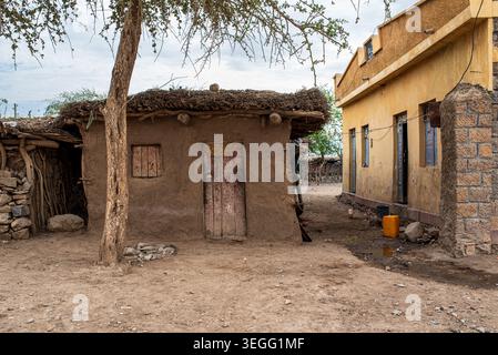 Une humble cabane de boue traditionnelle avec un toit de chaume se dresse à côté d'une maison moderne peinte en jaune dans un village éthiopien. Un jerrycan d'eau orange sur th Banque D'Images