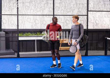 Divers amis masculins marchant court de padel bleu tenant des raquettes, des balles de tennis près d'une clôture en maille Banque D'Images