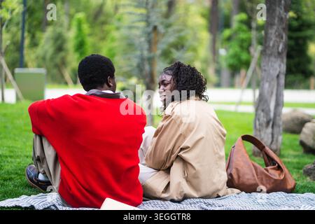 Heureux couple afro-américain amis proches se réunissant pour la date, assis sur l'herbe dans le parc et parlant. Banque D'Images