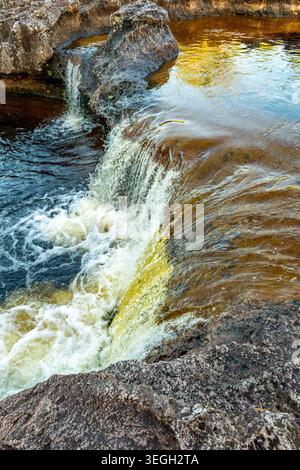 Une cascade descend en cascade des formations rocheuses, créant une mousse blanche dans des eaux tranquilles sous la lumière du soleil. Banque D'Images