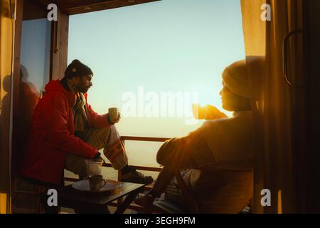 Un couple est assis sur un balcon en dégustant un thé chaud tout en regardant le lever du soleil doré sur le paysage de montagne à Chakrata, Uttarakhand, Inde. Banque D'Images