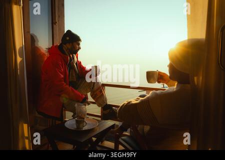 Un homme et une femme assis sur un balcon tenant des tasses de thé tout en profitant de la vue dorée du lever du soleil sur les collines de Chakrata, Uttarakhand, Inde. Banque D'Images