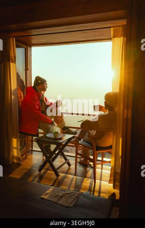 Un couple assis sur un balcon buvant du thé tout en regardant le lever du soleil sur les montagnes dans une atmosphère sereine à Chakrata, Uttarakhand, Inde. Banque D'Images