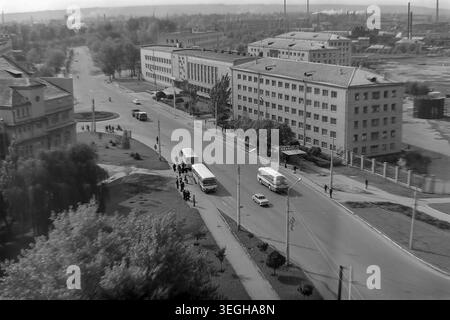 Sloviansk, fin des années 1970 - début des années 1980, rue Yunykh Komunariv (maintenant rue Bankivska). Une large perspective aérienne met en valeur une intersection de ville animée avec les transports publics soviétiques classiques. Plusieurs grands autobus de passagers sont arrêtés le long de la route principale près d'une aire d'attente désignée avec des piétons. Le paysage urbain est caractérisé par des bâtiments administratifs et résidentiels importants à plusieurs étages, des trottoirs soignés et des cheminées industrielles lointaines à l'horizon. Un merveilleux reflet de l'infrastructure civique et de la paix dans le Donbass. Banque D'Images