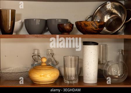 Une collection de divers ustensiles de cuisine, y compris des bols en bois, des gobelets en verre, des bouteilles et une passoire, est soigneusement stocké sur des étagères ouvertes rustiques. Banque D'Images