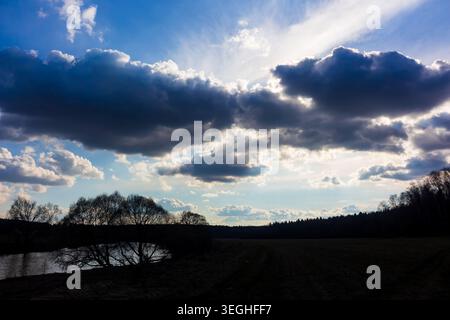 Des rayons de soleil spectaculaires traversent de lourds nuages orageux au-dessus d'une rivière tranquille et d'un paysage rural sombre, créant une atmosphère sombre au contraste saisissant Banque D'Images