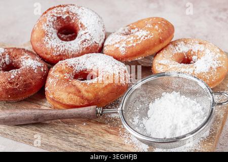 Une planche de bois contient plusieurs beignets fraîchement faits. Les beignets sont ronds et ont du sucre en poudre sur le dessus. Un petit tamis est à proximité pour ajouter du sucre Banque D'Images