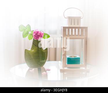 Une belle rose rose dans un petit vase en céramique verte se trouve sur une table ronde en verre à côté d'une lanterne vintage blanche avec une bougie turquoise brûlant à l'intérieur Banque D'Images