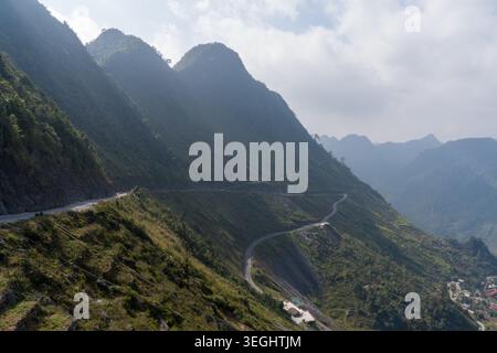 La route sinueuse traverse des pentes de montagne luxuriantes. Des collines verdoyantes s'étendent sous un ciel nuageux. Des pics accidentés s'élèvent au loin. Sentier courbe à travers des tanières Banque D'Images