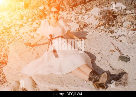 La mode de plage de femme se trouve sur de grandes pierres portant une élégante robe blanche et des lunettes de soleil pendant le coucher de soleil chaud de l'heure dorée Banque D'Images