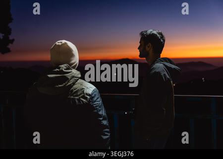 Deux aventuriers se tiennent sur une crête au crépuscule, admirant le coucher de soleil doré sur les collines et les vallées brumeuses à Chakrata, Uttarakhand, Inde. Banque D'Images
