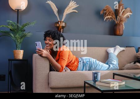 Femme afro-américaine allongée sur un canapé et utilisant son smartphone à la maison Banque D'Images
