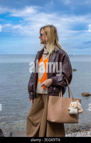 Femme blonde élégante debout par la côte de la mer calme dans une tenue d'automne décontractée et atmosphère paisible Banque D'Images