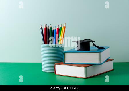 Une casquette de graduation miniature sur une pile de livres à côté d'un contenant de crayons de couleur. Obtention du diplôme, réussite scolaire, enseignement supérieur, lectures scolaires Banque D'Images