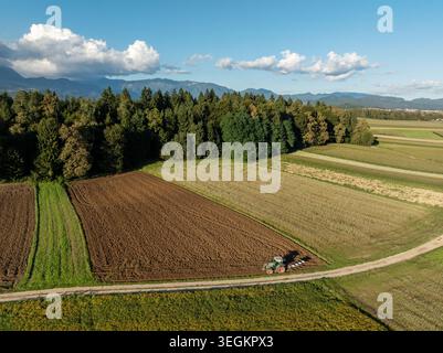Vue aérienne montre un tracteur travaillant sur des terres agricoles entourées d'arbres et de montagnes par temps clair dans la campagne. Banque D'Images