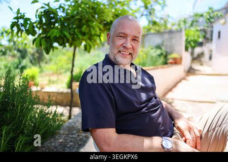 Homme mature souriant se relaxant en plein air dans le jardin méditerranéen ensoleillé, appréciant le style de vie paisible de vacances et l'atmosphère authentique de voyage d'été. Banque D'Images