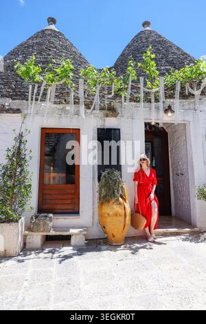 Femme élégante en robe rouge debout près de la maison traditionnelle de trulli à Alberobello, Italie pendant les vacances ensoleillées méditerranéennes. Banque D'Images