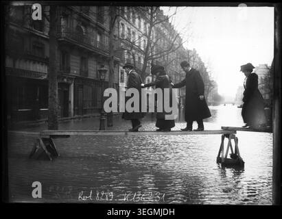 Les gens marchent prudemment sur des planches surélevées de l’avenue Ledru-Rollin dans le 12ème arrondissement de Paris lors des inondations du 30 janvier 1910, négociant les rues inondées et maintenant le mouvement le long des sentiers élevés au milieu de la montée des niveaux d’eau. Appuyez sur photo. Banque D'Images