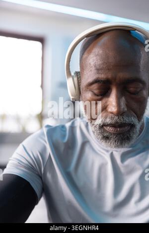 Homme afro-américain senior assis à la salle de gym portant un casque blanc et un manchon de compression noir Banque D'Images