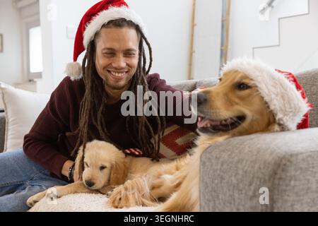 Homme souriant dans le pull marron Santa-Hat chiot caressant à la maison, retriever portant le chapeau de Père Noël près Banque D'Images