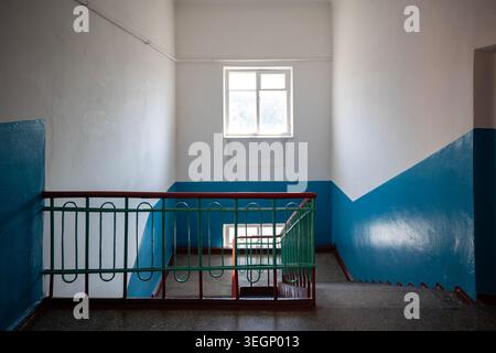 Une vue intérieure d'une cage d'escalier d'immeuble résidentiel avec des marches en béton, une main courante en métal vert, des murs peints en blanc et bleu, et une victoire Banque D'Images