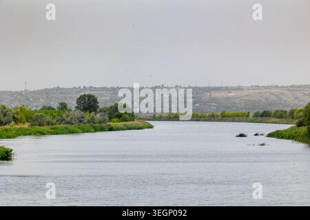 Une large rivière calme coule à travers un paysage vert rural avec des arbres et des roseaux poussant le long des rives sous un ciel gris nuageux avec des collines et des bâtiments Banque D'Images