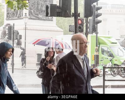 Londres, Royaume-Uni. 20 mai 2026, météo britannique. Un homme plus âgé en costume marche dans une averse près d'Admiralty Arch à la jonction du Mall et de Trafalgar Square, dans le centre de Londres, le 19 mai 2026, apparemment indemne par la pluie. Une femme avec un parapluie Union Jack passe derrière. Trafalgar sqaure est visible en arrière-plan. La figure composée au milieu de la douche soudaine du West End sur un jour de soleil et de douches à travers London.Credit:xiu bao/Alamy Live News Banque D'Images