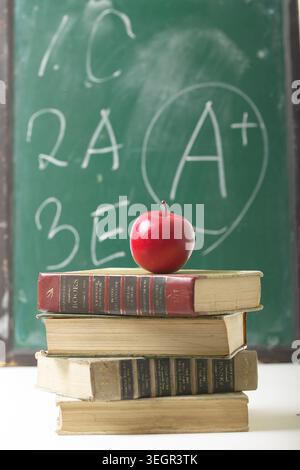 Une pomme rouge se trouve au sommet d'une pile de livres vintage sur un bureau encombré et à l'ancienne dans une salle de classe faiblement éclairée. Banque D'Images