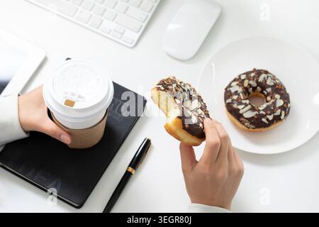 Une femme tient une tasse à café et un beignet au chocolat à son bureau, entourée d'un ordinateur portable, d'un cahier et d'un stylo. Banque D'Images