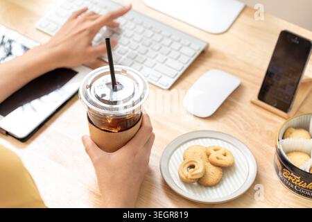 Une personne travaille sur un ordinateur portable dans un bureau moderne, sirotant du café glacé et profitant d'une assiette de biscuits. Banque D'Images