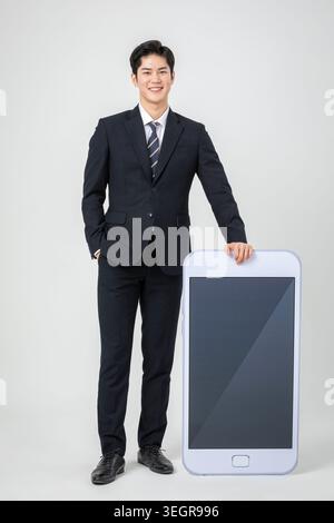 Un jeune homme asiatique en costume tient un smartphone géant, souriant. Banque D'Images