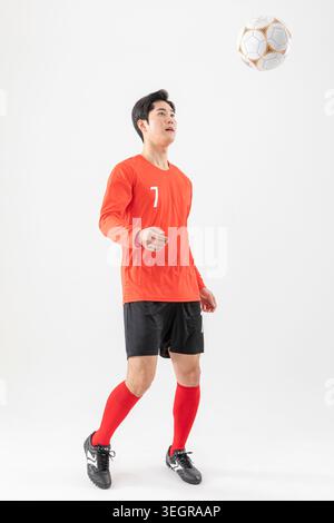 Un jeune homme asiatique en maillot de football, attrapant un ballon de football dans les airs. Banque D'Images