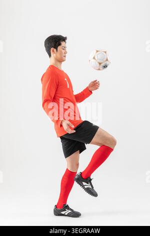 Un jeune asiatique portant un maillot rouge et un short noir jongle avec un ballon de football avec son pied gauche. Banque D'Images