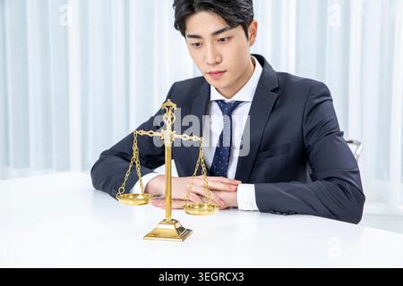 Un jeune homme d'affaires asiatique est assis à un bureau, regardant un ensemble de balances en laiton antiques. Banque D'Images